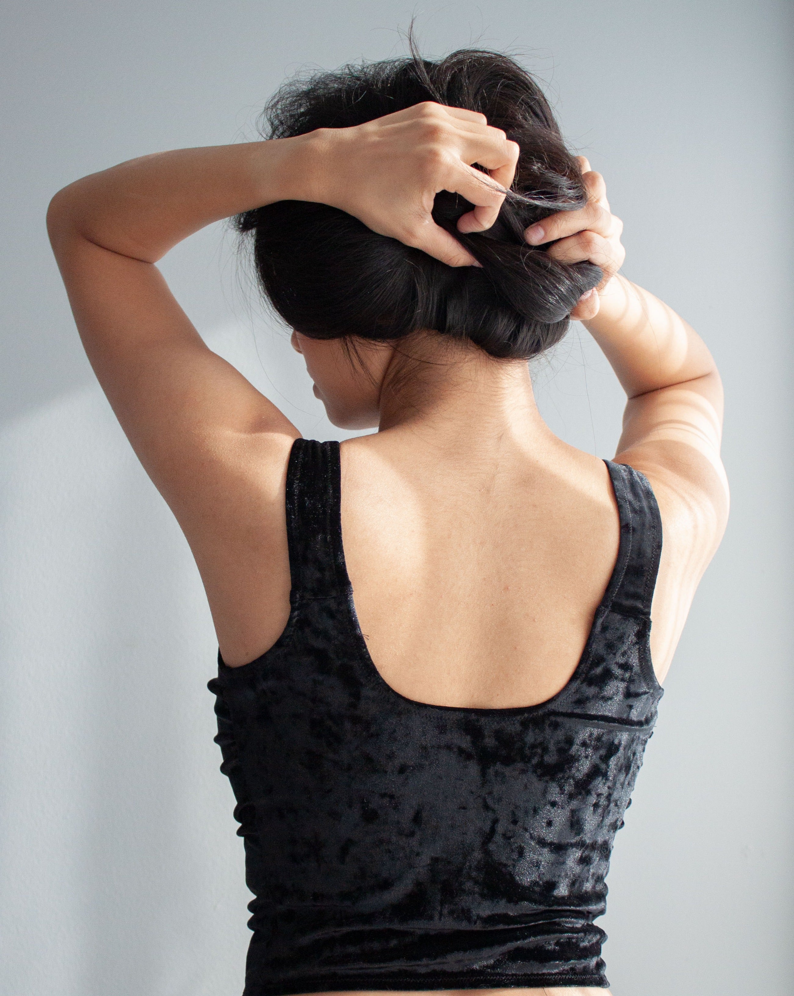 Model wearing a black crushed velvet custom-made corset top. Her back is facing the camera, she holds up her hair with both hands. The back of the corset has a scooped back with wide straps.