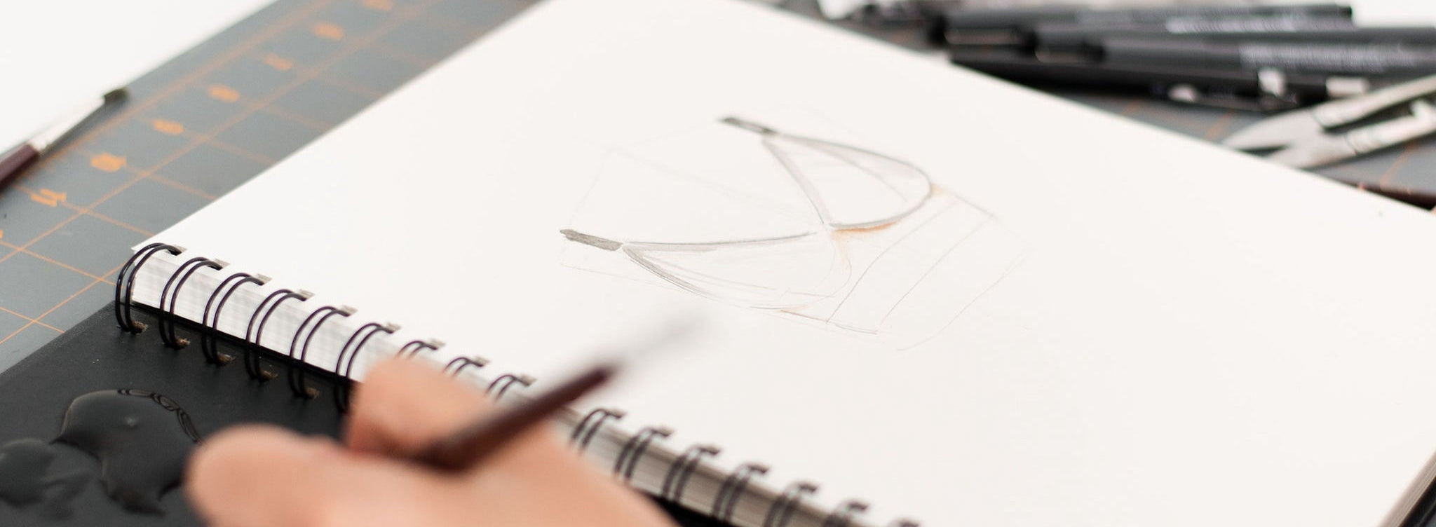 Hand holding a pen over a notebook with a drawing on a desk.