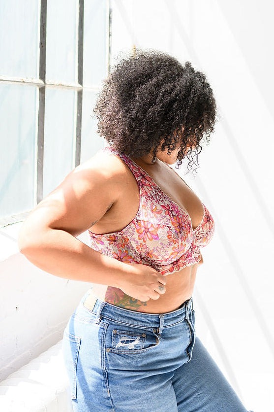 Woman in a floral lace bra and blue jeans standing in a bright room with large windows.