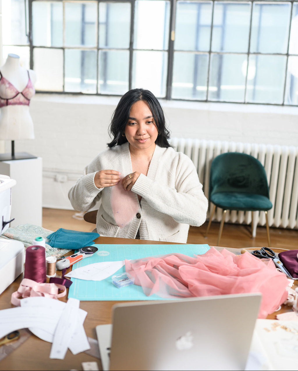 A woman sits at a desk in a bright sewing studio, working with pink fabric and sewing materials. A mannequin wearing a bra is visible in the background near large industrial windows.