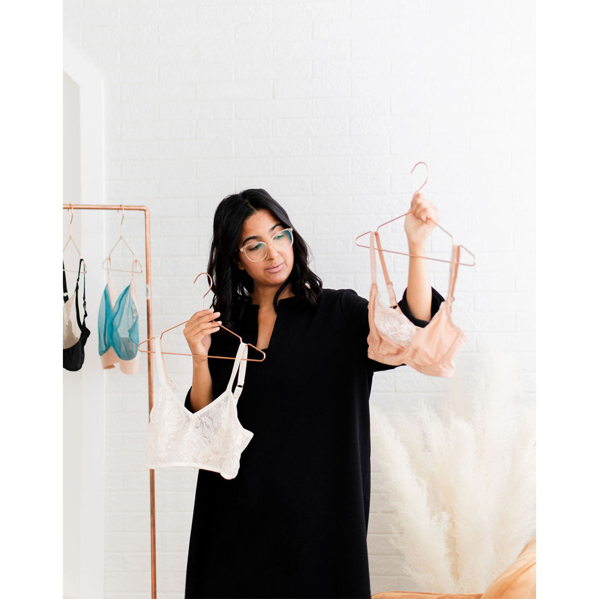 Rubies Bras founder holding up two bras and showing to client during a virtual fitting. She has brown skin with black medium hair wearing black. The bras she is holding are copper lace and ivory lace.