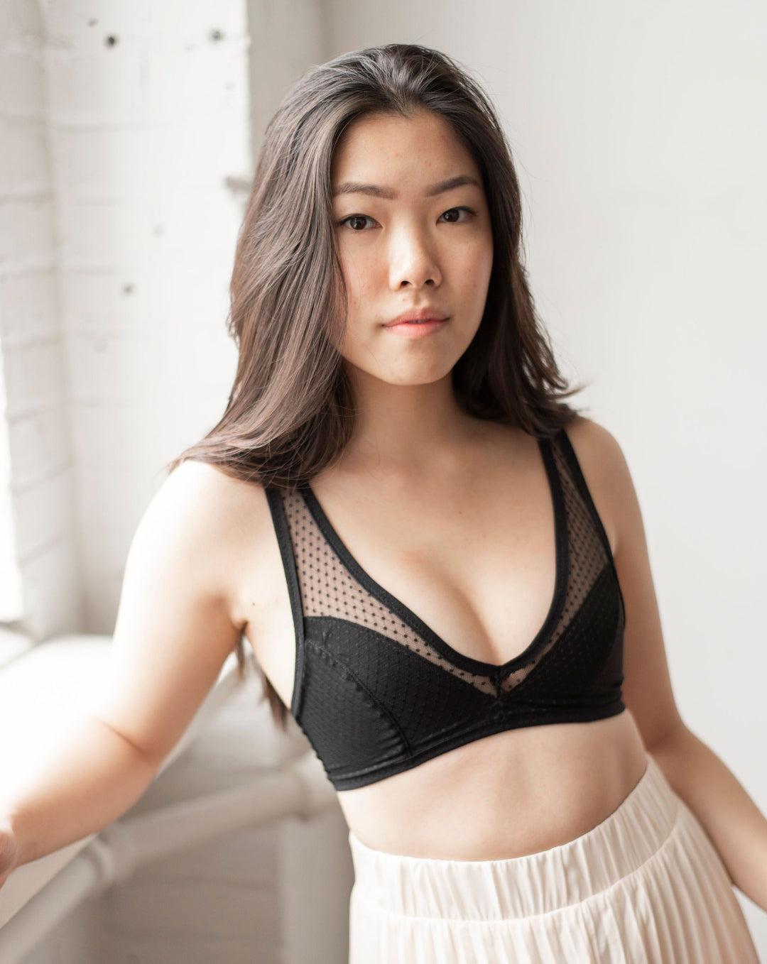 Rubies Bras. Female with long black hair wearing a black dotted lace wirefree bra with deep plunge v neckline. Front facing half body shot on white brick background.