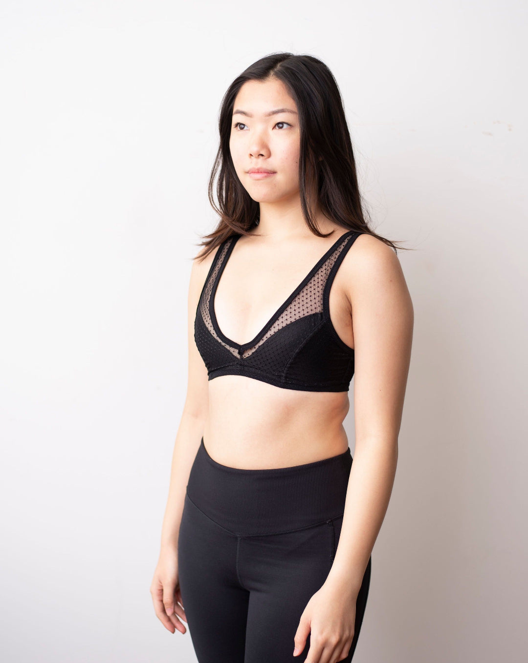 Rubies Bras. Female with long black hair wearing a black dotted lace wirefree bra with deep plunge v neckline. Side facing full body shot on white background.