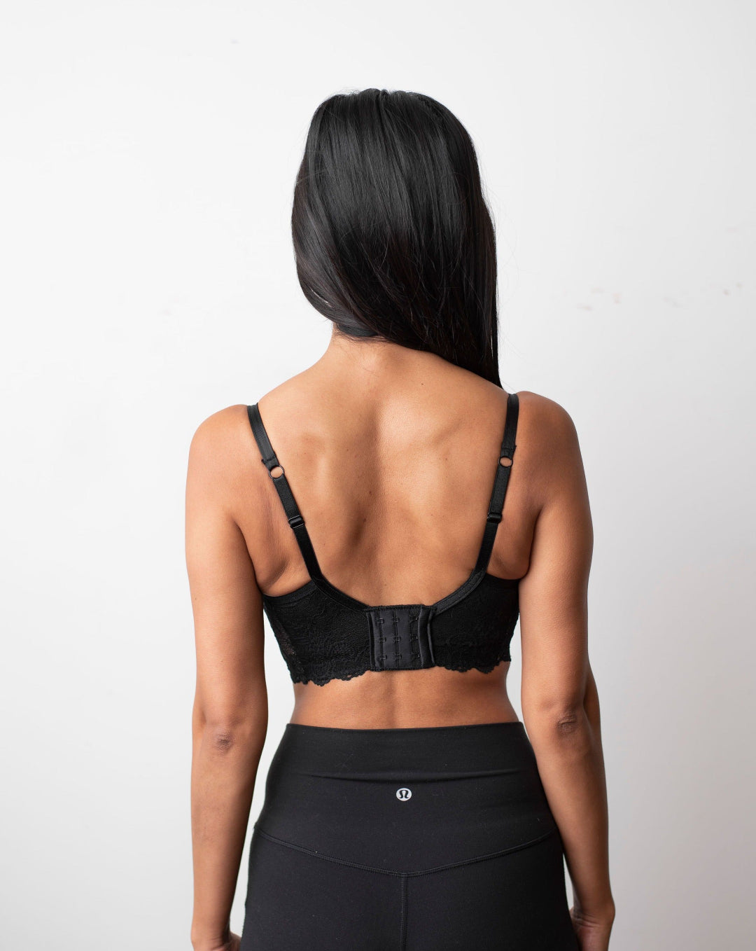 Ruhee with Rubies Bras, a female with long black hair wearing a black scalloped lace wirefree bra with deep plunge v neckline. Full back body shot on white back drop .