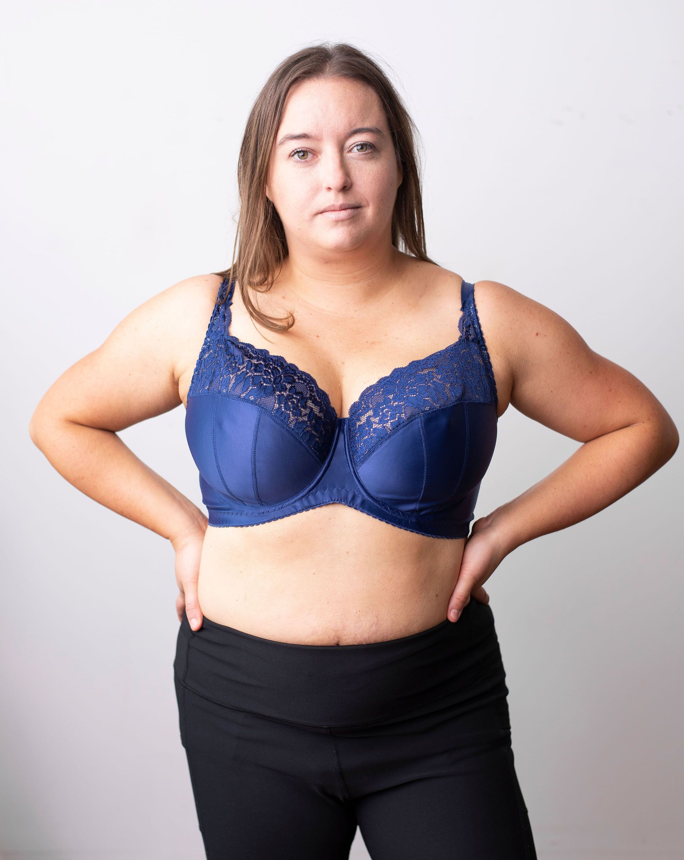 Model with straight brown hair stands with her hands on her waist. She wears a high coverage wired, blue bra. Half body frontal shot in front of a plain white background.