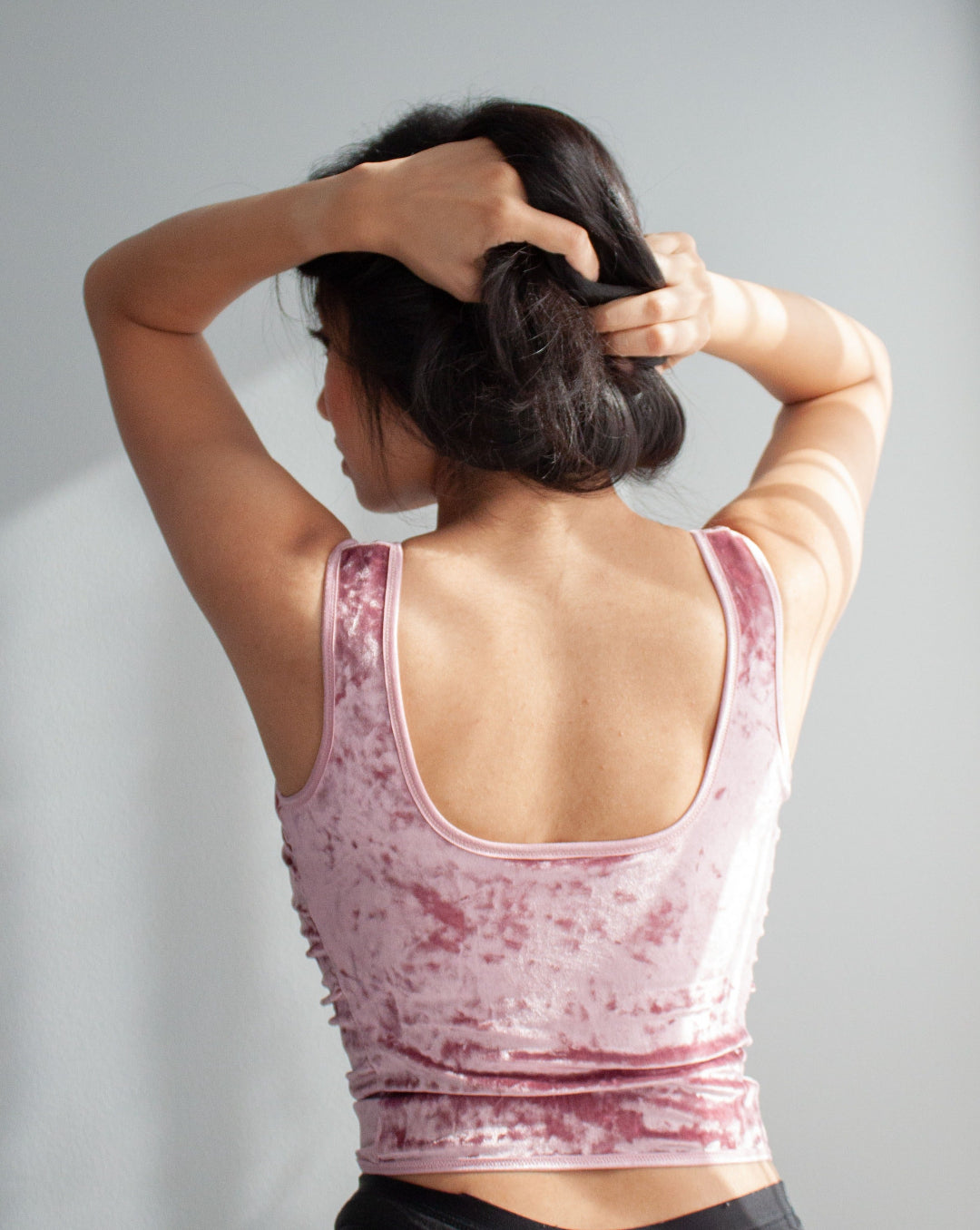 Model wearing a baby pink crushed velvet custom-made corset top. Her back is facing the camera, she holds up her hair with both hands. The back of the corset has a scooped back with wide straps. 