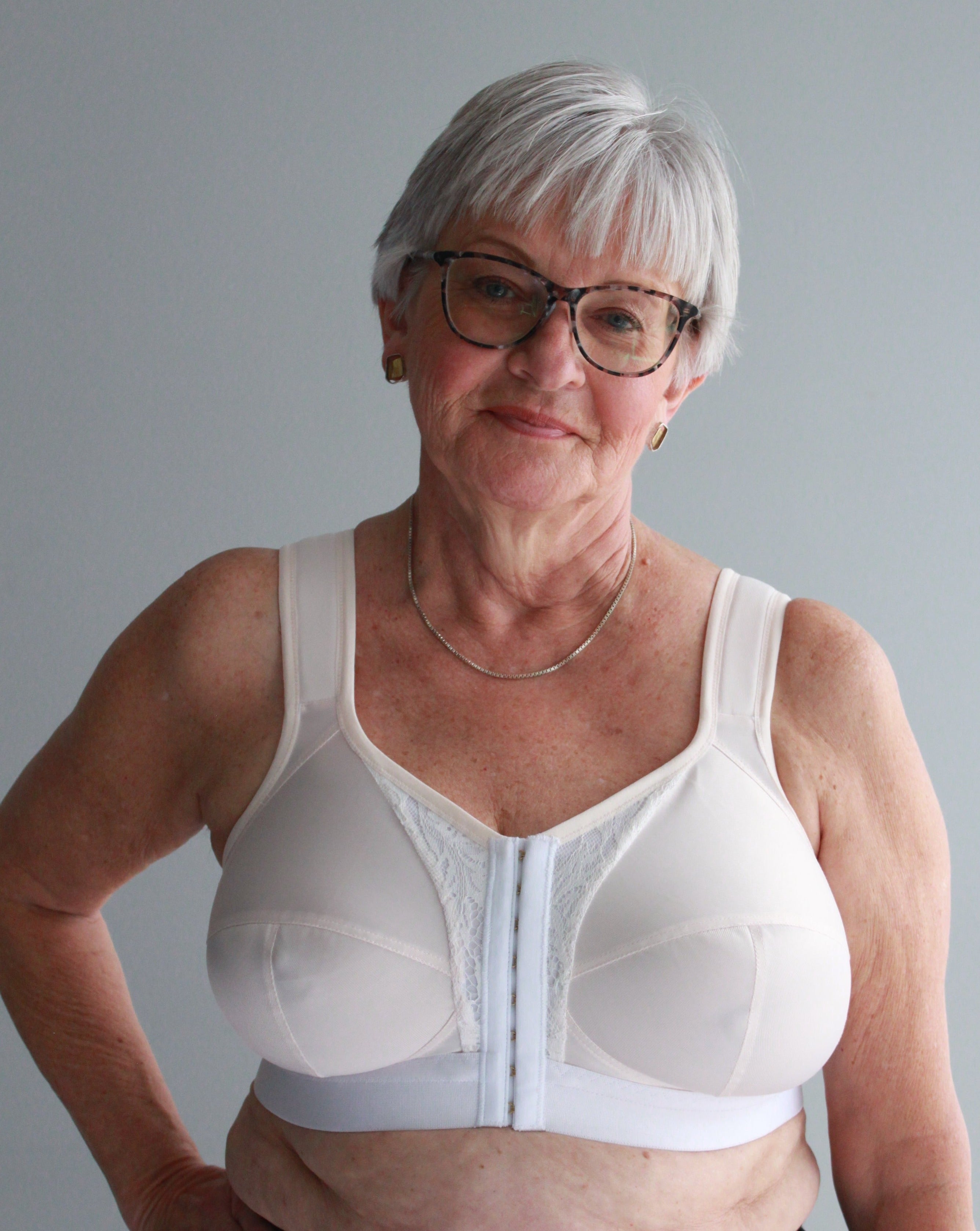 A woman with short grey hair wearing a white, full coverage, wired Ingrid Bra from Rubies Custom Bras on a grey backdrop.
