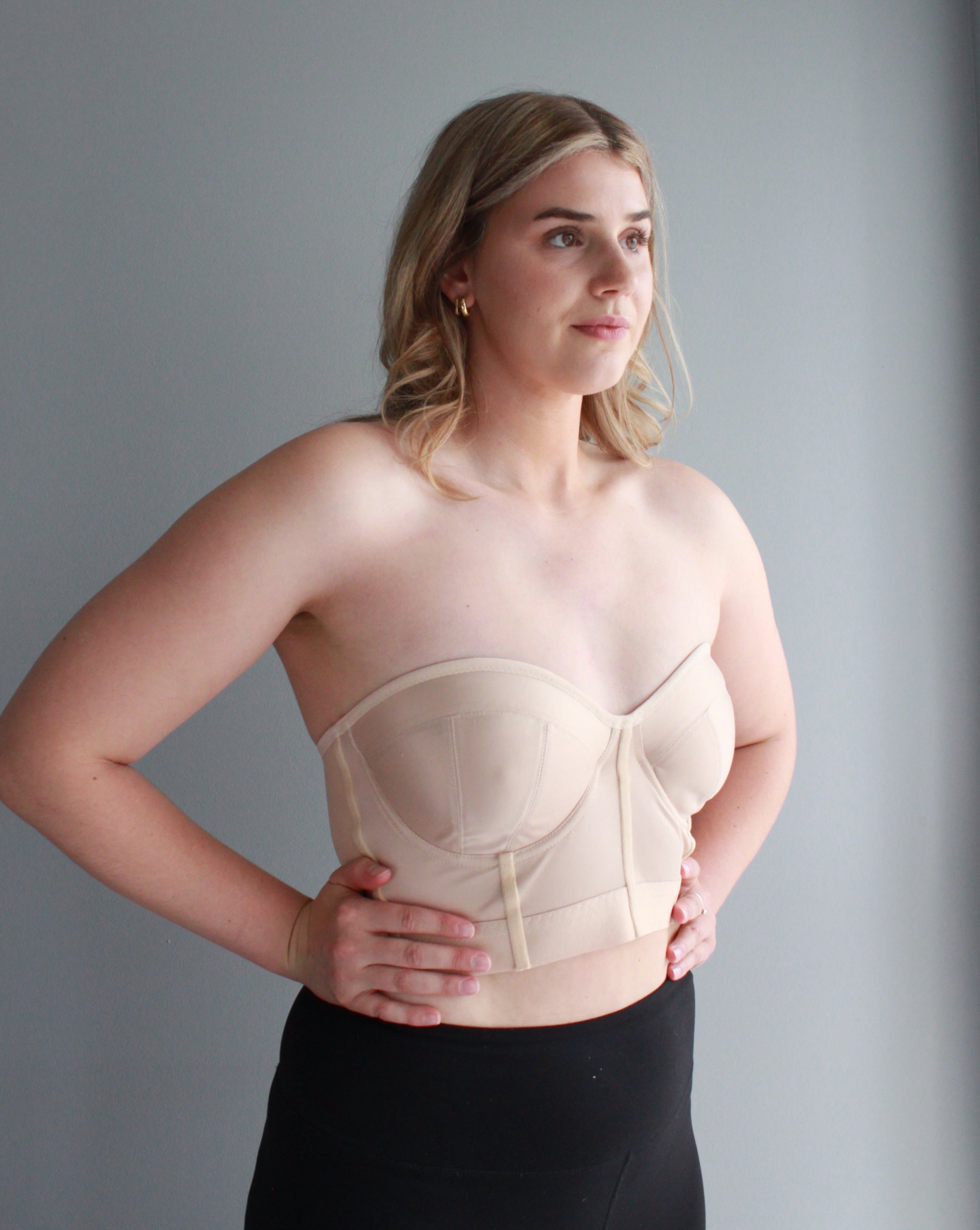 A model with short blond hard with her hands on her hips wearing a strapless beige bustier from Rubies Custom Bras. 