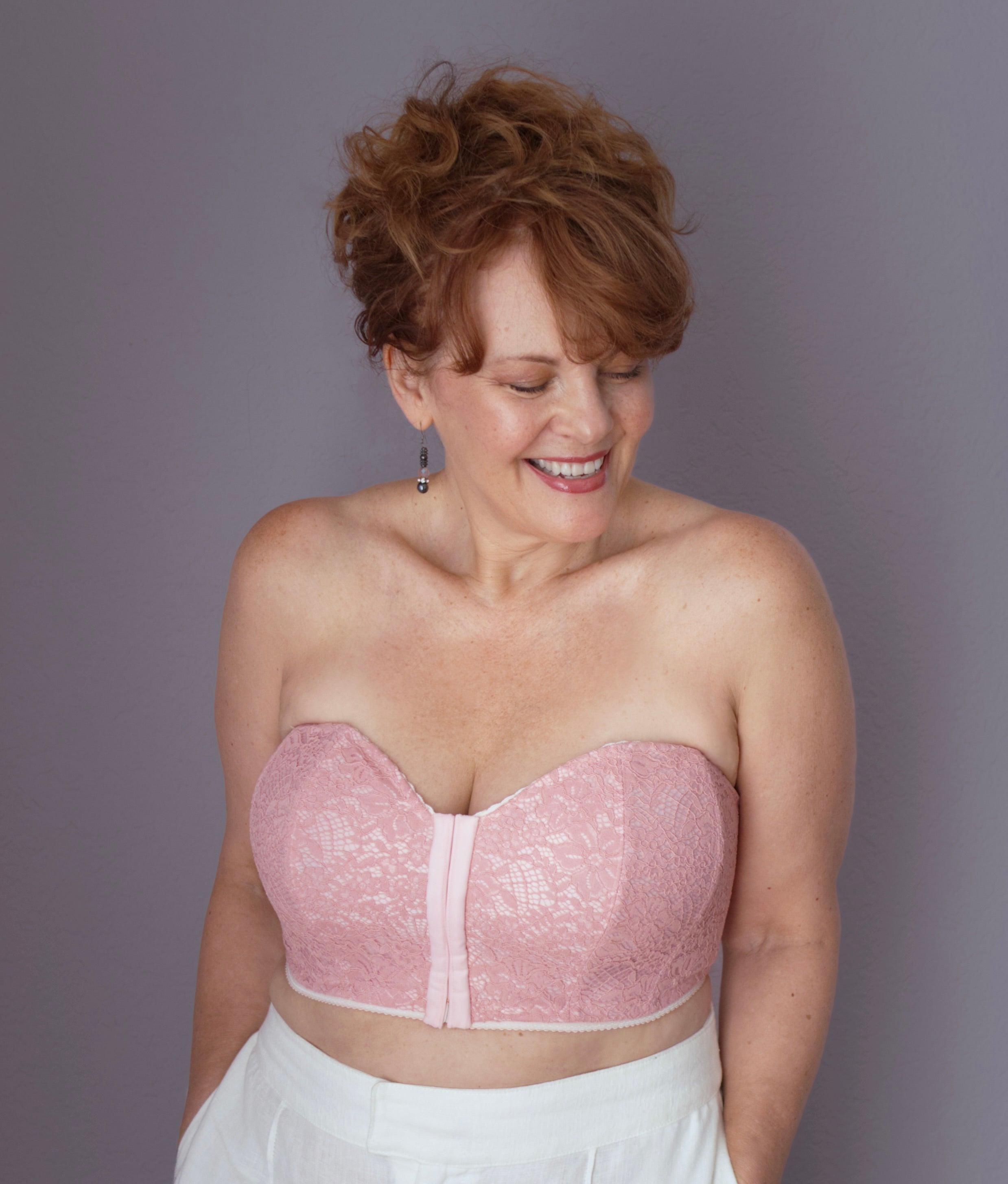 A model with curly red hair smiling to the side with her hands in her pockets wearing a strapless bustier in pink from Rubies Custom Bras