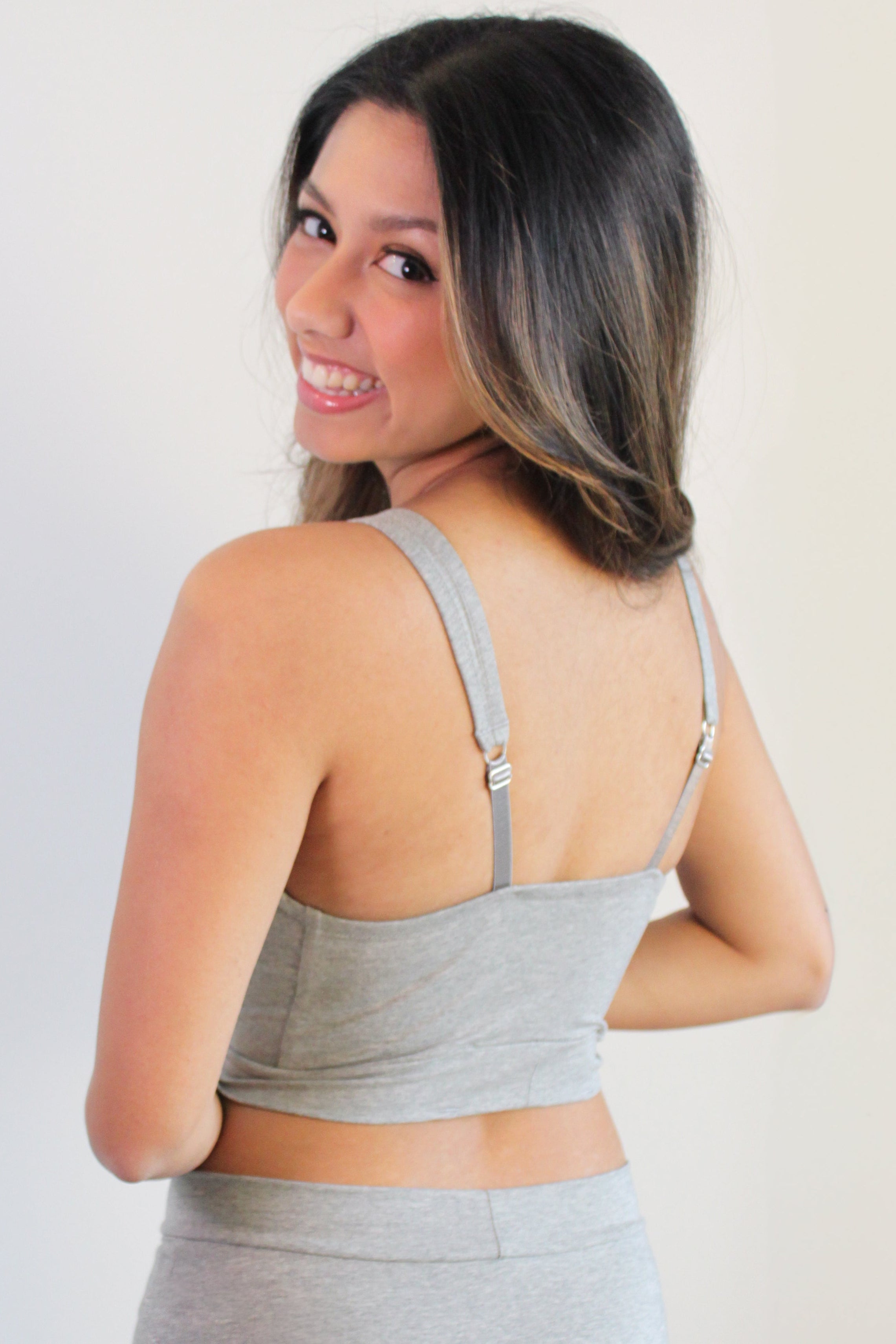 A model with long dark hair, with her back turned to the camera while smiling wearing the Sahaara Lounge bralette from Rubies Custom Bras in the colour grey.