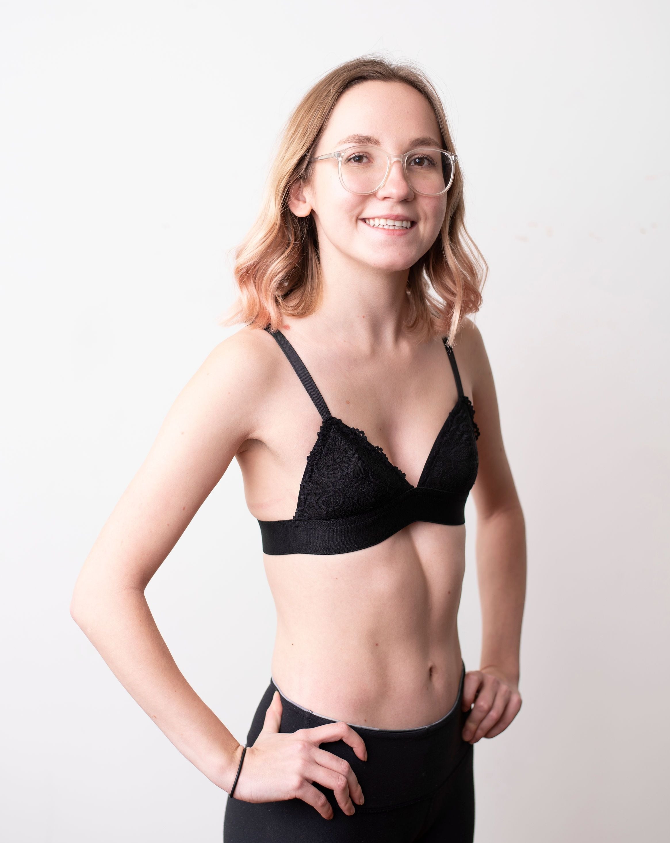 Haley from Rubies Bras, a female with medium blond hair wearing a black scalloped lace wirefree bra from our petites collection. Full side body shot on white back drop.