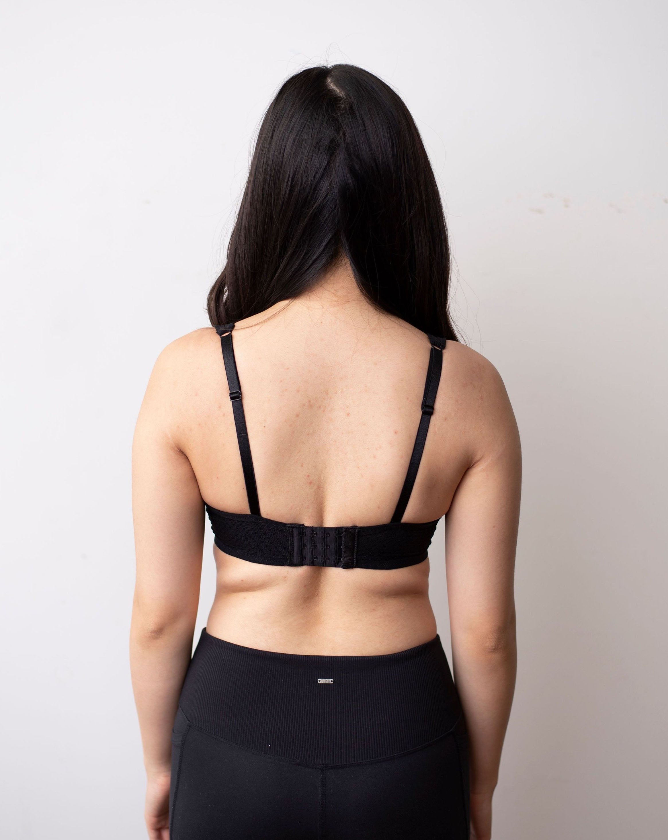 Half body back shot of female with long back hair against a white backdrop. Wearing a black, dotted, plunge bra.