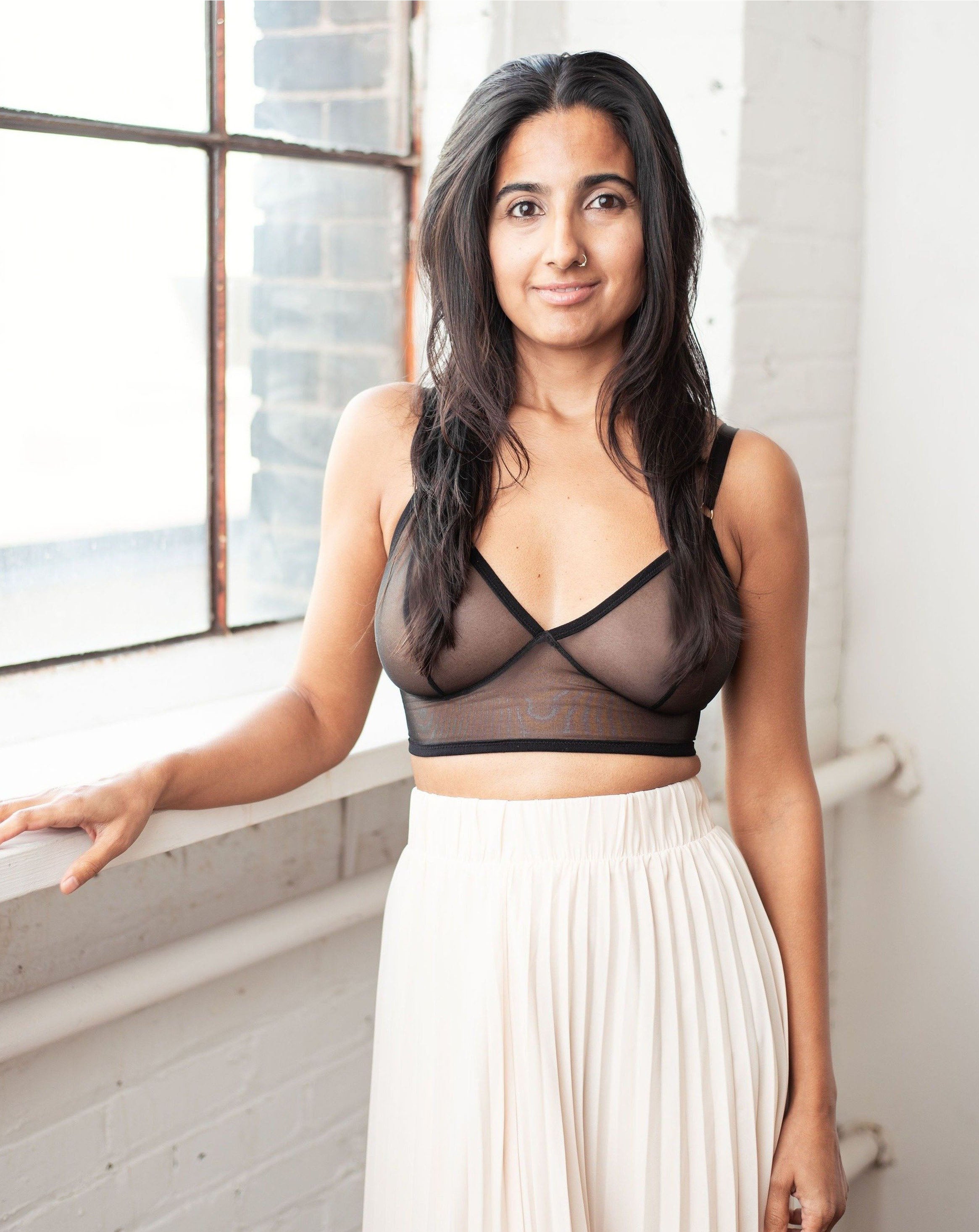 Ruhee with Rubies Bras, a female with long black hair wearing a black sheer wirefree bra with a low v neckline. Full front body shot on white brick back drop with a window.