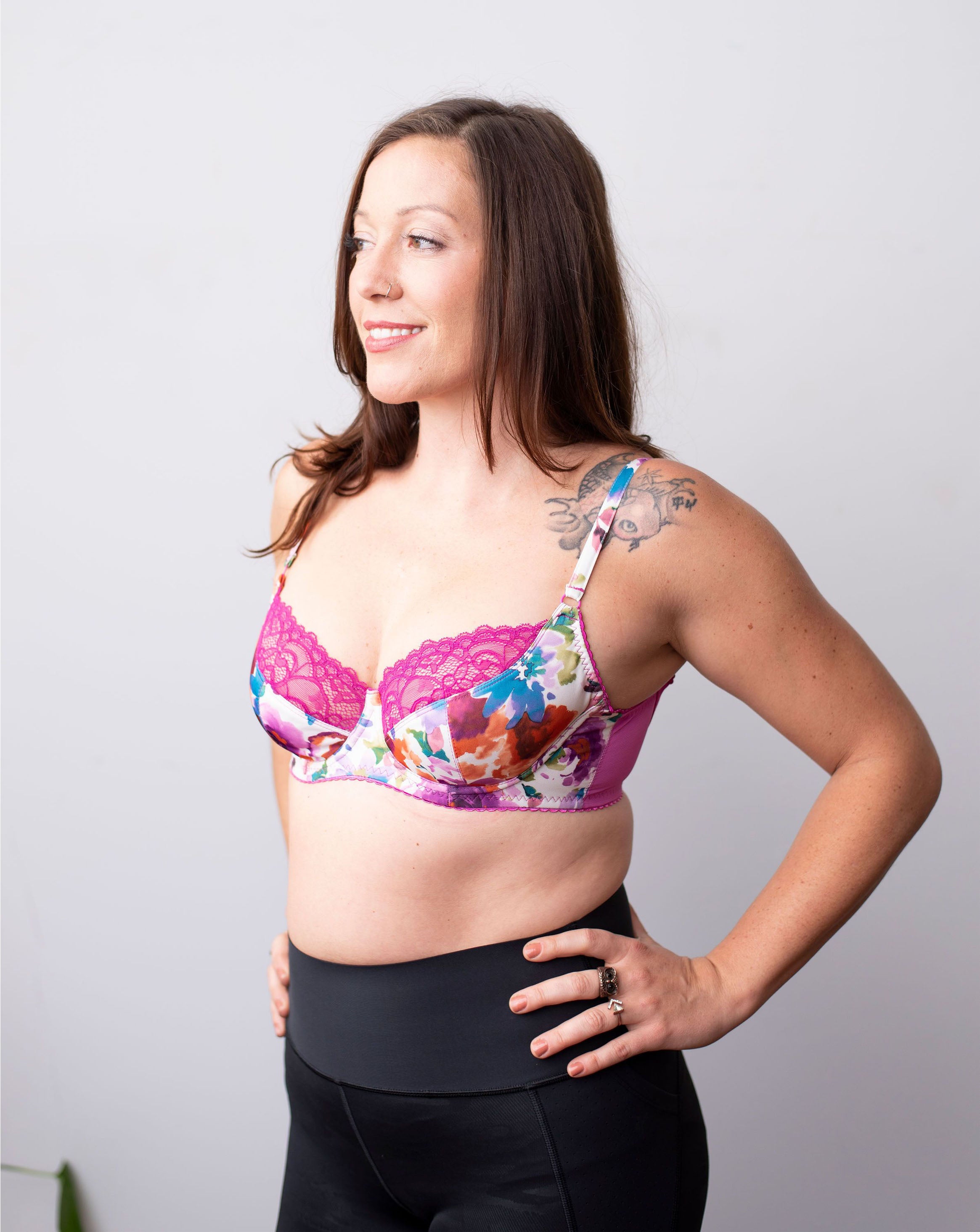 Model with straight brown hair stands with her hands on her waist. She wears a wired floral pink bra with hot pink lace. Half body frontal shot in front of a plain white background.