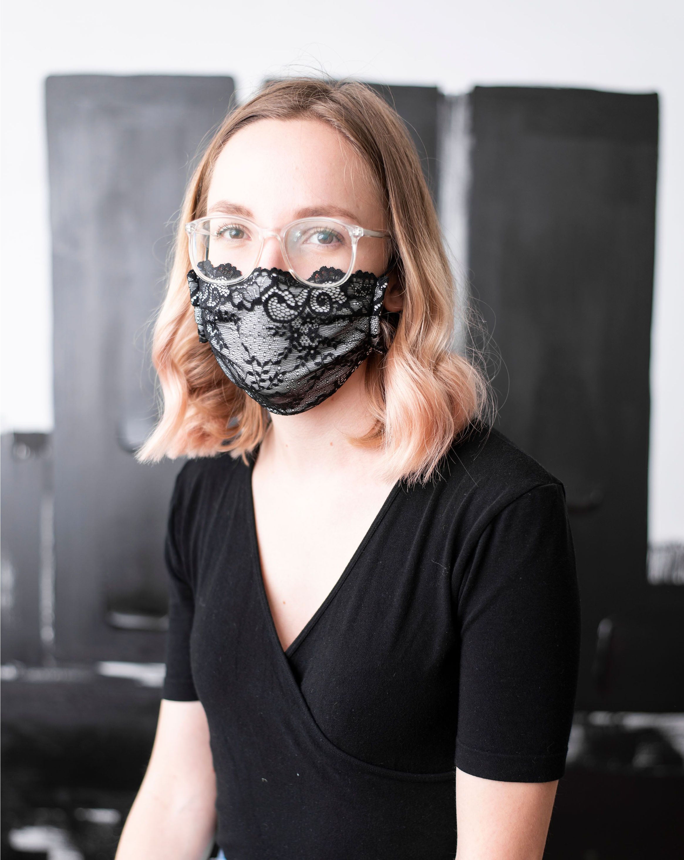 Model with brown hair and clear frame glasses wearing a black lace, organic cotton face mask. Front shot of the upper body.