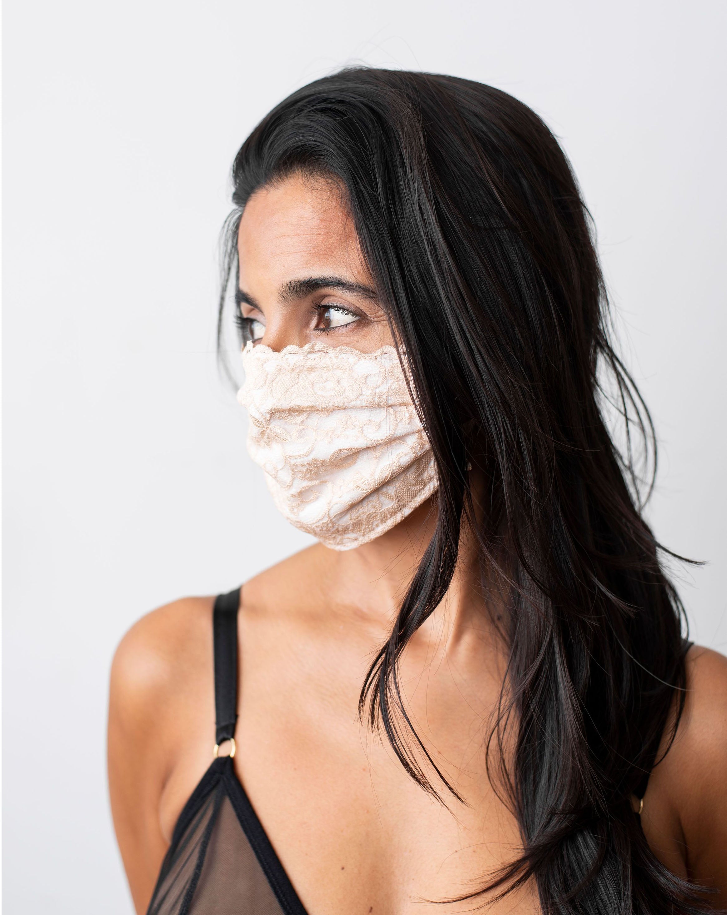 Ruhee, female with long black hair, is wearing a beige lace, organic cotton facemask. Side shot against a plain white backdrop.